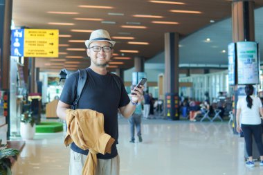 Happy young Asian man with a backpack is holding jacket and smartphone while smiling to the camera, blurry airport lobby background.