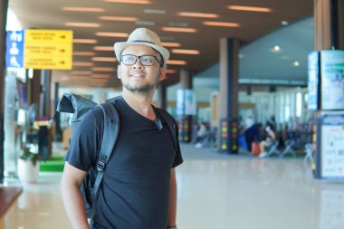 Joyful young Asian man wearing straw hat and backpack to go on a holiday, waiting and looking around the airport lounge.