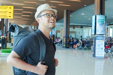 Excited young Asian man wearing straw hat and backpack to go on a holiday, looking around the airport lounge.
