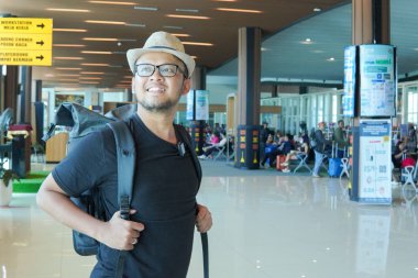 Excited young Asian man wearing straw hat and backpack to go on a holiday, looking around the airport lounge.