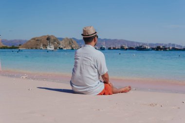 Back view of a young Asian man wearing beach hat sitting and enjoying the pink beach scenery in Labuan Bajo.
