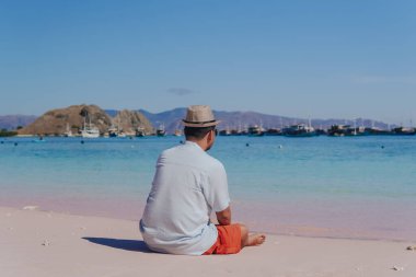 Back view of a young Asian man wearing beach hat sitting and enjoying the pink beach scenery in Labuan Bajo.