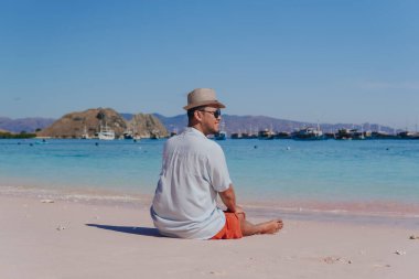 Back view of a young Asian man wearing beach hat sitting and enjoying the pink beach scenery in Labuan Bajo.