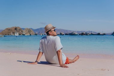 Back view of a young Asian man wearing beach hat sitting and enjoying the pink beach scenery in Labuan Bajo.