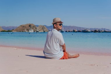 Back view of a young Asian man wearing beach hat sitting and enjoying the pink beach scenery in Labuan Bajo.