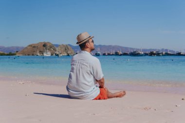 Back view of a young Asian man wearing beach hat sitting and enjoying the pink beach scenery in Labuan Bajo.