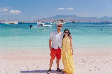 Joyful young Asian couple standing and posing to take picture at Pink Beach, Labuan Bajo, with ocean and boats sailing on the background.