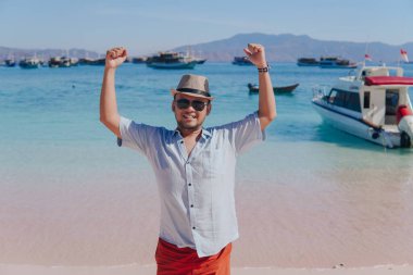 Excited young Asian man wearing shirt and beach hat posing on Pink Beach, Labuan Bajo with his arms spreaded.