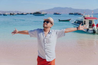 Excited young Asian man wearing shirt and beach hat posing on Pink Beach, Labuan Bajo with his arms spreaded.