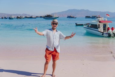 Excited young Asian man wearing shirt and beach hat posing on Pink Beach, Labuan Bajo with his arms spreaded.