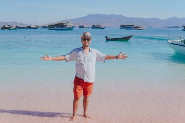 Excited young Asian man wearing shirt and beach hat posing on Pink Beach, Labuan Bajo with his arms spreaded.