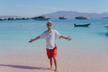Excited young Asian man wearing shirt and beach hat posing on Pink Beach, Labuan Bajo with his arms spreaded.
