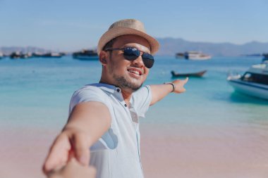 Cheerful young Asian man pointing to the scenery on Pink Beach, Labuan Bajo while holding hands with a woman.
