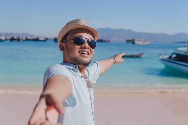 Cheerful young Asian man pointing to the scenery on Pink Beach, Labuan Bajo while holding hands with a woman.