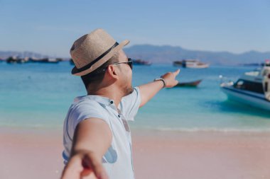 Cheerful young Asian man pointing to the scenery on Pink Beach, Labuan Bajo while holding hands with a woman.