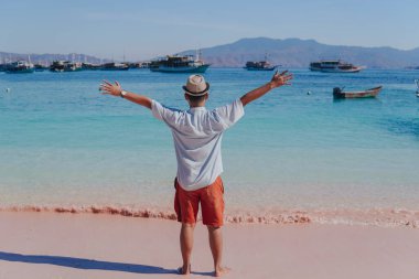 Back view of young Asian man wearing shirt and beach hat posing with his arms spreaded on Pink Beach, Labuan Bajo.