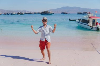 Joyful young Asian man wearing beach hat feeling excited while jumping in the air on Pink Beach, Labuan Bajo.