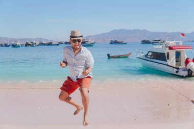 Joyful young Asian man wearing beach hat feeling excited while jumping in the air on Pink Beach, Labuan Bajo.
