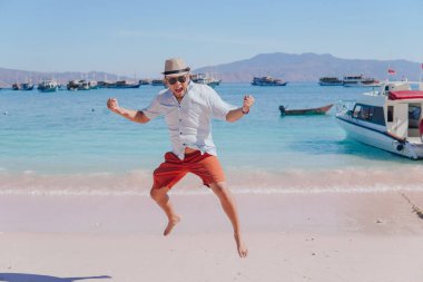 Joyful young Asian man wearing beach hat feeling excited while jumping in the air on Pink Beach, Labuan Bajo.