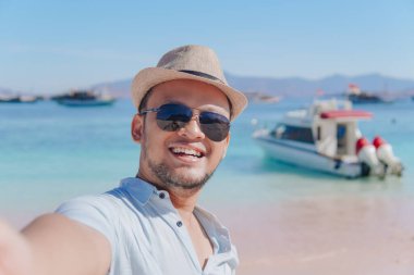 Cheerful young Asian man smiling while taking selfie picture at Pink Beach, Labuan Bajo.