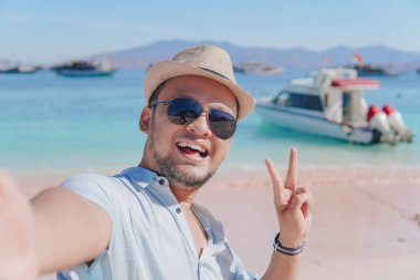 Happy young Asian man wearing shirt and hat is doing peace finger gesture while taking selfie picture at Pink Beach, Labuan Bajo.