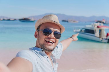 Excited young Asian man taking selfie picture while pointing to the scenery on Pink Beach, Labuan Bajo.