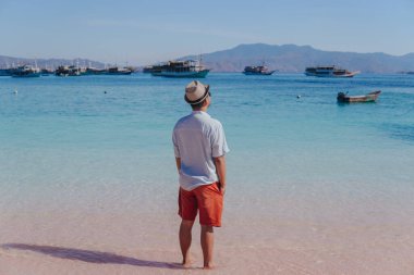 Back view of young Asian man wearing shirt and beach hat enjoying scenery at Pink Beach, Labuan Bajo.