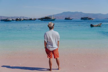 Back view of young Asian man wearing shirt and beach hat enjoying scenery at Pink Beach, Labuan Bajo.