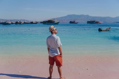 Back view of young Asian man wearing shirt and beach hat enjoying scenery at Pink Beach, Labuan Bajo.