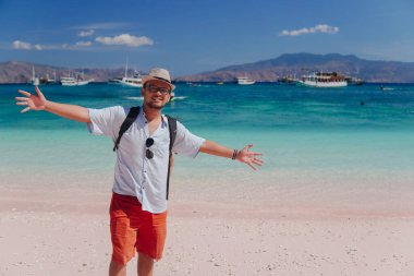 Cheerful young Asian man bringing backpack posing with his arms spread at Pink Beach, Labuan Bajo.