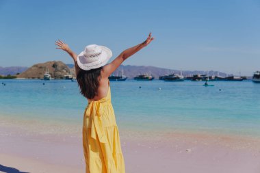 Back view of an excited young Asian woman wearing yellow dress standing and posing on a beach with her arms raised.