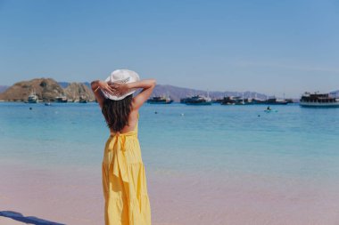 Back view of a carefree young Asian woman wearing yellow dress standing and enjoying the pink beach scenery in Labuan Bajo.