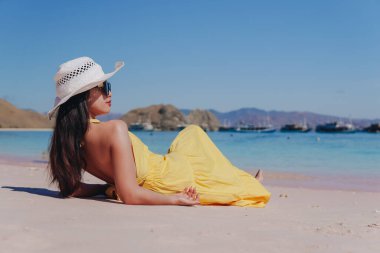 Back view of a young Asian woman wearing yellow dress sitting and enjoying the pink beach scenery in Labuan Bajo.
