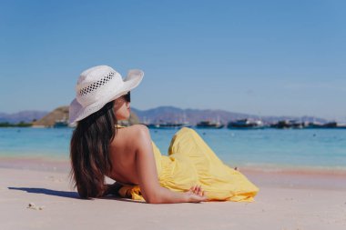 Back view of a young Asian woman wearing yellow dress sitting and enjoying the pink beach scenery in Labuan Bajo.