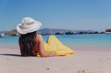 Back view of a young Asian woman wearing yellow dress sitting and enjoying the pink beach scenery in Labuan Bajo.