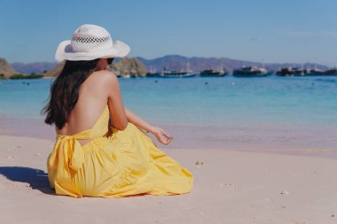 Back view of a young Asian woman wearing yellow dress sitting and enjoying the pink beach scenery in Labuan Bajo.