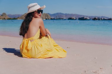 Back view of a young Asian woman wearing yellow dress sitting and enjoying the pink beach scenery in Labuan Bajo.