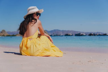 Back view of a young Asian woman wearing yellow dress sitting and enjoying the pink beach scenery in Labuan Bajo.