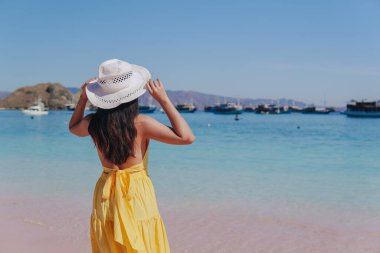 Back view of a carefree young Asian woman wearing yellow dress standing and enjoying the pink beach scenery in Labuan Bajo.