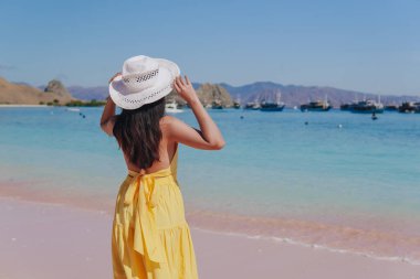 Back view of a carefree young Asian woman wearing yellow dress standing and enjoying the pink beach scenery in Labuan Bajo.