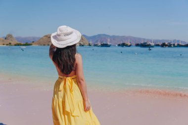 Back view of a carefree young Asian woman wearing yellow dress standing and enjoying the pink beach scenery in Labuan Bajo.