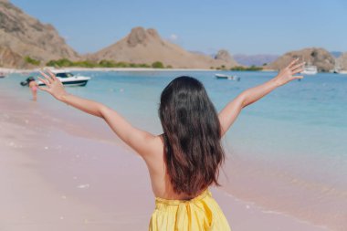Back view of an excited young Asian woman wearing yellow dress standing and posing on a beach with her arms raised.