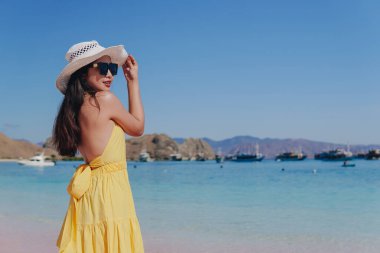 Back view of a carefree young Asian woman wearing yellow dress standing and enjoying the pink beach scenery in Labuan Bajo.