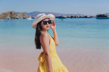 Back view of a carefree young Asian woman wearing yellow dress standing and enjoying the pink beach scenery in Labuan Bajo.