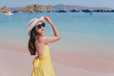 Back view of a carefree young Asian woman wearing yellow dress standing and enjoying the pink beach scenery in Labuan Bajo.