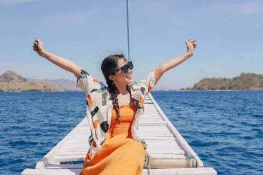 Gülümseyen Asyalı genç kadın heyecanla kollarını açıyor. Okyanusta bir teknede Labuan Bajo manzarasının tadını çıkarıyor..