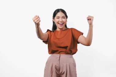 Kahverengi gömlekli neşeli genç Asyalı kadın yumruklarını mutlu bir ifadeyle kaldırıyor, beyaz arka planla izole edilmiş..