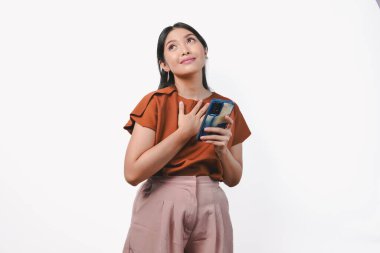 Mutlu, düşünceli, genç Asyalı kadın gülümseyen, elleri göğsünde, izole edilmiş beyaz arka plan üzerinde telefonu tutan..