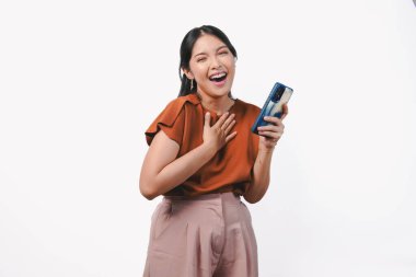 Mutlu, düşünceli, genç Asyalı kadın gülümseyen, elleri göğsünde, izole edilmiş beyaz arka plan üzerinde telefonu tutan..