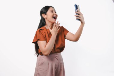 Kahverengi tişörtlü gülümseyen Asyalı kadın elinde akıllı bir telefonla beyaz arka planda yalnız poz veriyor..